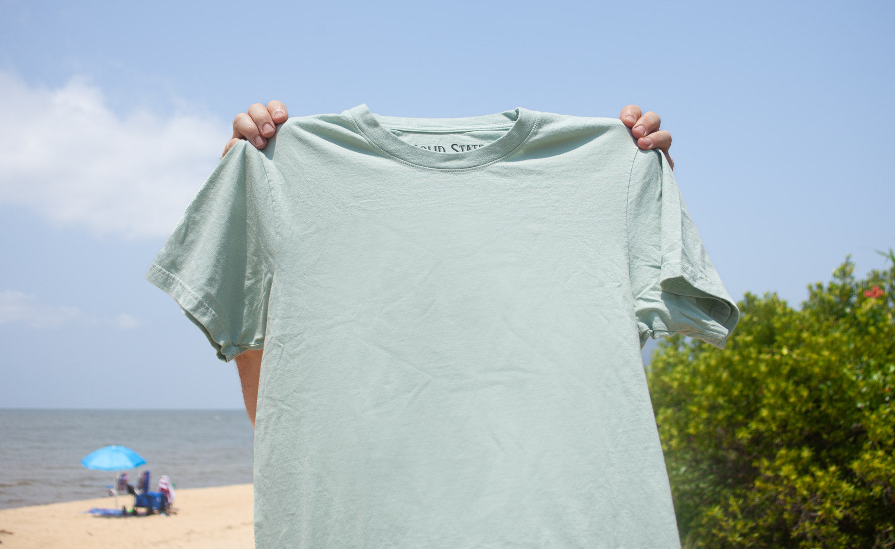 A sage green made in usa t-shirt being held up by a person on a beach