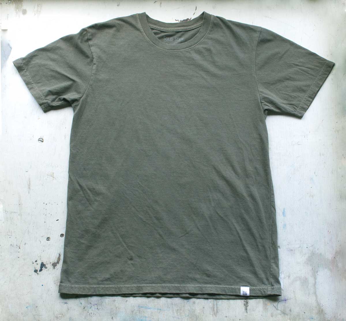 A Solid State Clothing Natural Dye t-shirt lies against a white background. The color of the shirt is Basil, which is a darker, muted shade of green