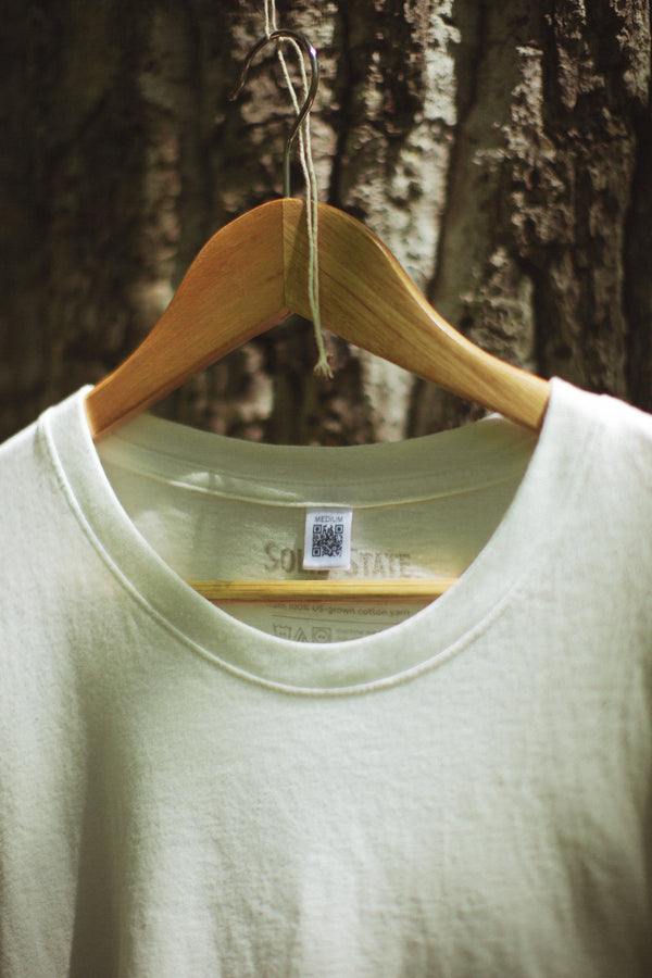 A white Solid State Clothing t-shirt is shown on a hanger. There is a tag with a QR code, which shows customers how the t-shirt is entirely made in the USA and tracks every step of the process behind how the shirt is made